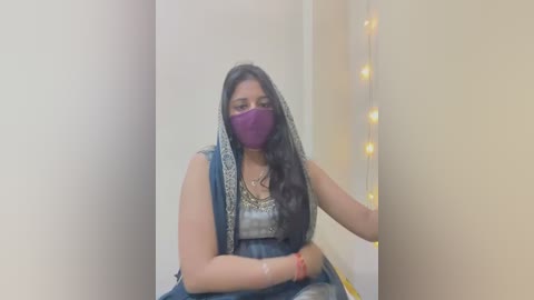 Media: Video of a South Asian woman with long black hair, wearing a purple mask and blue saree, seated indoors beside a string of warm fairy lights.