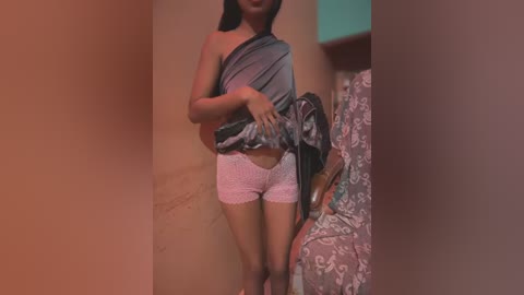 Media: Video of a young woman with medium brown skin and long black hair, standing in a dimly lit hallway. She wears a gray tank top and pink lace shorts, holding a black jacket. Background features beige walls and a floral-patterned armchair.