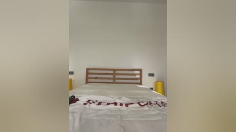 Media: A video of a minimalist bedroom with a wooden headboard, white bed linens, and a red message written in Japanese on the bedspread. The room has plain white walls and two yellow cylindrical nightstands.