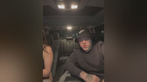 Media: Video of a man in a black hoodie, sunglasses, and a black hat seated in a dimly lit car, next to a topless woman with long hair, visible tattoo on his forearm.