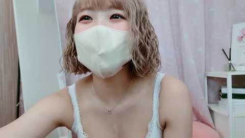 Media: Video of a young Asian woman with shoulder-length, wavy blonde hair, wearing a white face mask, a white lace camisole, and a delicate necklace. She's indoors, with pastel-colored curtains and a wooden shelf in the background.