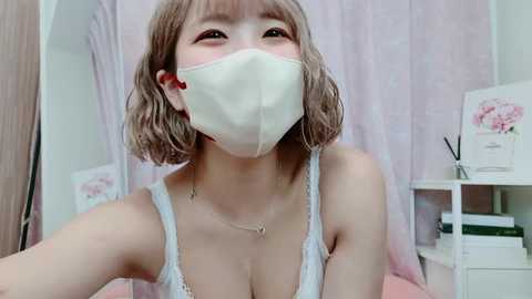 Video of a young Asian woman with short, wavy blonde hair, wearing a white face mask, a lacy white bra, and a delicate necklace. She is indoors, with pastel pink curtains and a white shelf in the background.