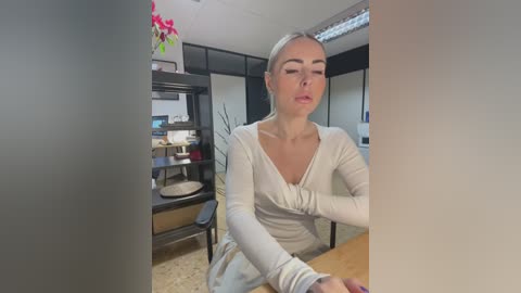 Media: Video of a blonde woman with fair skin, wearing a white dress, sitting at a wooden desk, eyes closed, in a modern office with shelves, a plant, and a bright ceiling light.