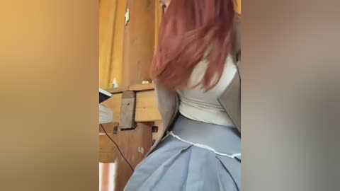 Media: Video of a person with long, reddish-brown hair, wearing a white top and blue skirt, standing in a wooden room with a plugged-in electronic device on a table.