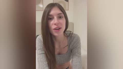 Media: A video of a young Caucasian woman with long brown hair, wearing a grey crop top, smiling, standing in a minimalist hallway.