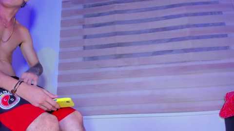 Media: Video of a shirtless man with tattoos, wearing red shorts and holding a yellow Nintendo Switch, sitting in front of a wooden-paneled window.