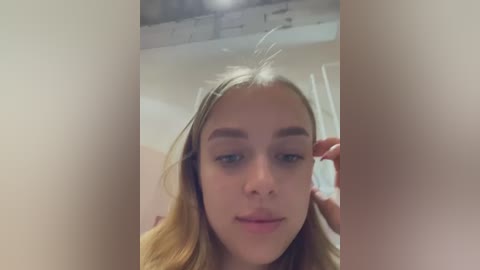 Media: A close-up video of a young Caucasian woman with blonde hair, fair skin, and blue eyes, wearing minimal makeup. She is indoors, possibly in a bathroom, with blurred, neutral-colored walls in the background.