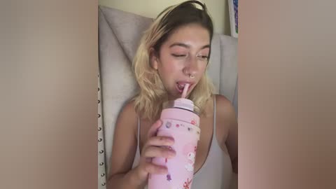 Media: Video of a young woman with light skin and shoulder-length blonde hair, wearing a pink tank top, drinking from a pink, floral-patterned sippy cup, lying on a beige couch with a textured headrest.