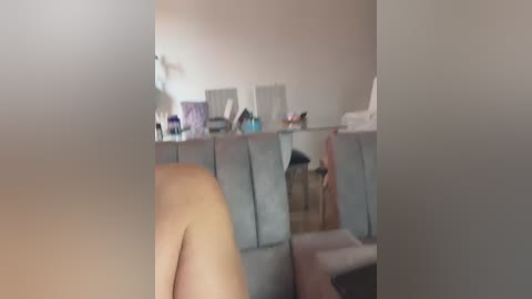 Media: A blurry video of a nude person sitting on a grey couch, with a white wall and a cluttered shelf in the background, featuring various items and a white towel.