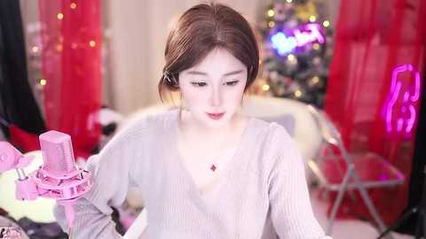 Media: A video of a young East Asian woman with fair skin, straight brown hair, and a soft, light grey V-neck sweater, seated at a pink vanity table with a gold necklace. Background features a festive room with a decorated Christmas tree and red curtains.