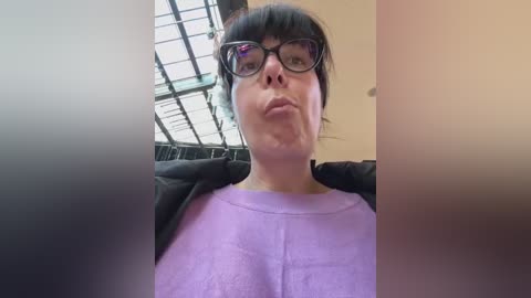 Media: A video of a woman with short black hair, wearing black-rimmed glasses and a purple t-shirt, puckering her lips, taken indoors with a high ceiling and large windows in the background.