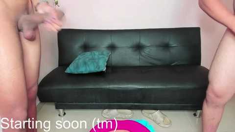 Media: Video of a black leather couch with teal pillow, surrounded by two nude men, one on each side, with erect penises. Background: light pink wall, white floor, scattered clothes. Text: \"Starting soon (m)\