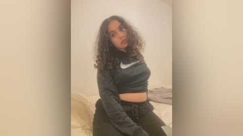 Media: Video of a young woman with medium skin tone and curly hair, wearing a dark blue crop top and black pants, sitting on a bed with beige sheets and pillows, in a simple, dimly-lit room.