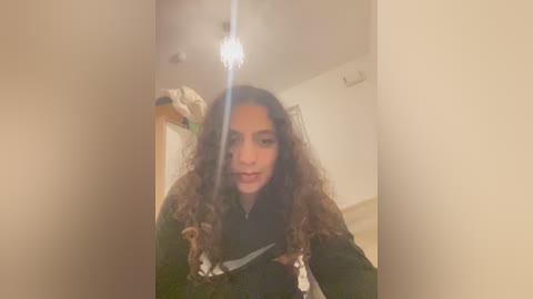 Media: A video of a young woman with long, curly brown hair, wearing a black jacket, standing in a dimly lit hallway with beige walls, a hanging light fixture, and a closet visible in the background.