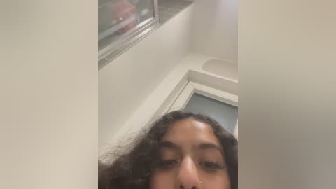 Media: Video of a young woman with medium brown skin and curly dark hair, taken from a low angle. She is wearing glasses and a nose ring. Background includes a window with a red car outside, a white door, and a beige wall.