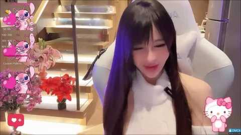 Media: A digital video of a young Asian woman with long black hair, wearing a white top, sitting in a plush chair in a modern room with a glass staircase and pink flowers.