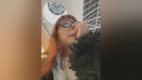 Media: Video of a young woman with long red hair and glasses, wearing a white top, standing in a modern building with a circular skylight and metal staircase.