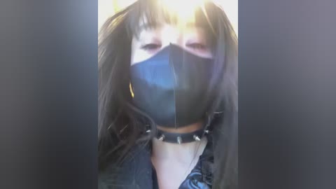 A video of a person with long, dark hair, wearing a black face mask and a spiked choker, standing in a dimly lit room.