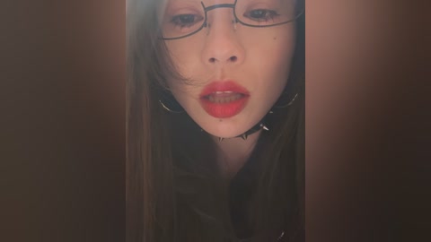 Video of a young woman with fair skin, dark hair, and glasses, wearing a black choker and red lipstick, set against a dark, blurred background.