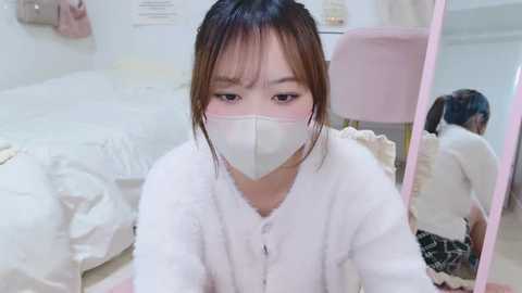 Media: A video of a young Asian woman with straight black hair, wearing a white face mask and white fuzzy robe, sitting on a bed in a softly lit, minimalist bedroom.