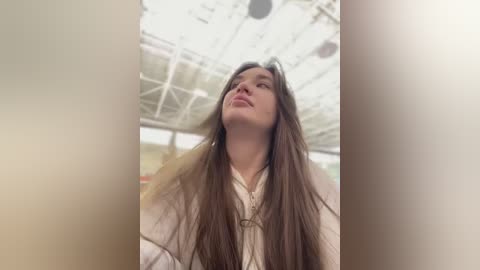 Media: Video of a young woman with long, straight brown hair, wearing a white shirt, looking upward with a contemplative expression. Background features a modern, glass-roofed structure with metal beams.