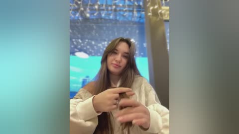 Media: Video of a young woman with long brown hair, wearing a cream-colored sweater, gesturing with both hands in front of a blue and gold-themed backdrop, likely in a festive or celebratory setting.