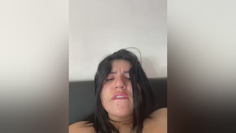 Media: Video of an Asian woman with shoulder-length black hair, closed eyes, and a distressed expression, sitting on a black leather couch against a white wall.