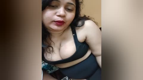 Media: Video of a plus-sized woman with fair skin, wearing a black bra and black high-waisted pants, posing seductively, with a neutral background and a hint of a colorful patterned fabric.