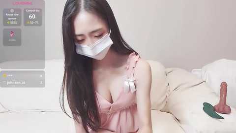 Media: Video of a young East Asian woman with long black hair, wearing a pink camisole and white face mask, lying on a beige bed, with a sex toy beside her.