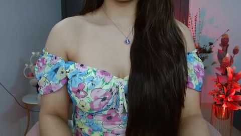 Media: Video of a young woman with long dark hair, wearing a colorful floral off-shoulder dress. She's indoors with a floral wallpaper background, a vase of red flowers, and a small table visible.