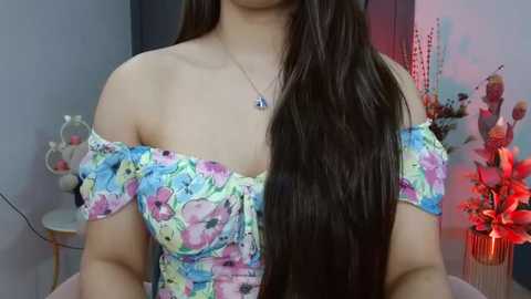 Media: Video of a light-skinned woman with long, straight dark brown hair, wearing a colorful, off-the-shoulder floral dress, with a silver necklace. Background features a gray wall, red floral arrangement, and a white chair.