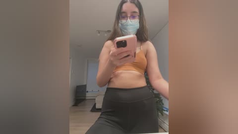 Media: Video of a young woman with light brown skin, wearing a face mask, glasses, and an orange sports bra, taking a selfie in a modern, minimalist apartment.