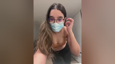 Media: Video of a young woman with long, wavy brown hair, wearing glasses, a light blue mask, and a beige tank top, adjusting her glasses in a dimly lit room.