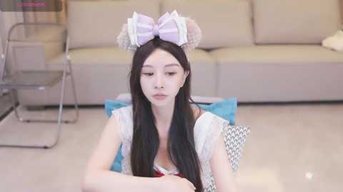 Media: A video of a young East Asian woman with long black hair, wearing a white lace top and a large pastel bow headpiece. She sits on a blue cushion in a modern living room with beige furniture.