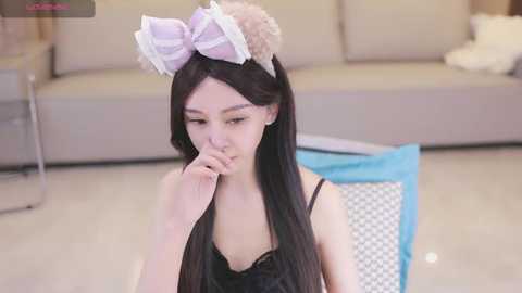 Media: A young Asian woman with long black hair, wearing a black lace top, sits on a blue cushion, looking contemplative with a finger to her mouth. She has a large, pink and white bow headband. The background features a beige sofa and a gray wall.