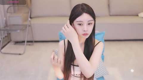 Media: Video of a young Asian woman with long black hair, sitting on a blue cushion, fixing her hair in a modern, minimalistic living room with beige couches and glass coffee table.