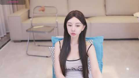 Media: Video of an East Asian woman with long black hair, fair skin, wearing a gray tank top, seated on a blue cushion in a modern living room with beige sofa, glass table, and metal chair.