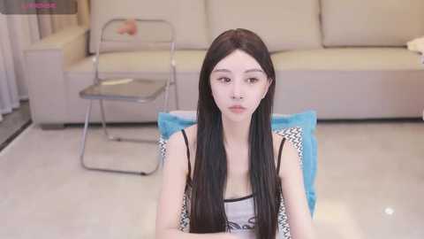 Media: Video of a young East Asian woman with long, straight black hair, wearing a sleeveless black and white top, sitting on a blue mat in a modern, minimalist living room with beige couch and metal chair.