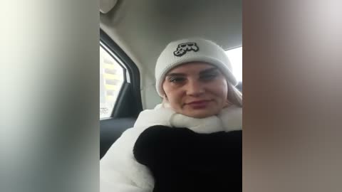 Media: Video of a Caucasian woman with light skin, wearing a white beanie with an \"Ewok\" logo, a white scarf, and a black and white sweater, sitting in a car.