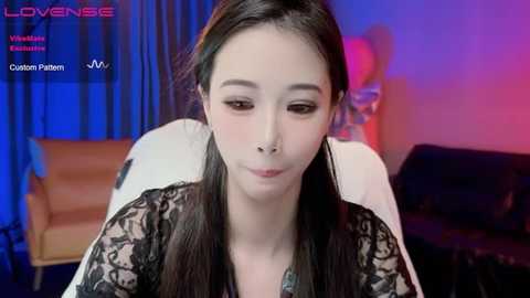 Media: Video of a young Asian woman with long black hair, wearing a black lace top, seated indoors with blue and red lighting.