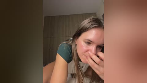 Media: A video captures a young woman with long, light brown hair, wearing a teal t-shirt, hiding her face with her hand, in a dimly-lit bedroom with beige curtains and a closet.