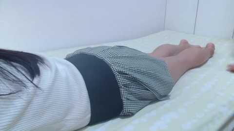 Media: Video of a woman lying face down on a bed, wearing a white ribbed top and a black-and-white checkered skirt. The room has white walls and a polka-dotted bedspread.