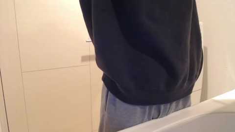 Media: Video of a person from the back, wearing a black hoodie and light blue sweatpants, standing in a bathroom with beige tiled walls and a white sink.