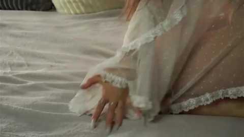 Media: Video of a woman in a sheer, white lace gown with intricate detailing, lying on a bed with light-colored sheets. Her hand, adorned with long, manicured nails, gently touches the bed. The setting appears intimate and serene.