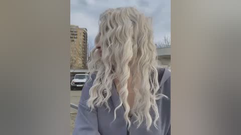 Media: A video of a woman with voluminous, wavy, platinum blonde hair, wearing a light gray sweater, standing outdoors beside a modern high-rise building and parked cars.