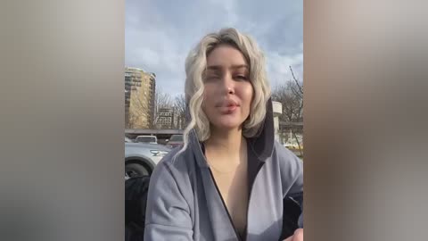 Media: Video of a young woman with light skin and wavy, platinum blonde hair, wearing a gray jacket, standing in an urban setting with tall buildings and parked cars in the background.