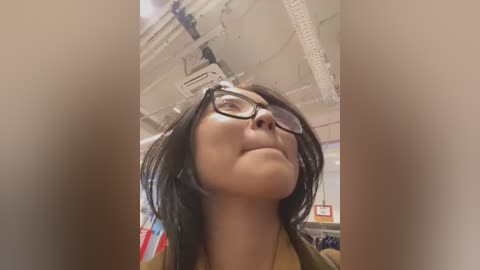 Media: Video of a young woman with fair skin and dark hair, wearing glasses, looking up, in a classroom with fluorescent lights and an American flag.