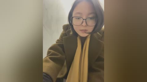 Media: Video of an East Asian woman with short, straight brown hair, wearing glasses and a brown coat, leaning against a beige wall, slightly blurred background.