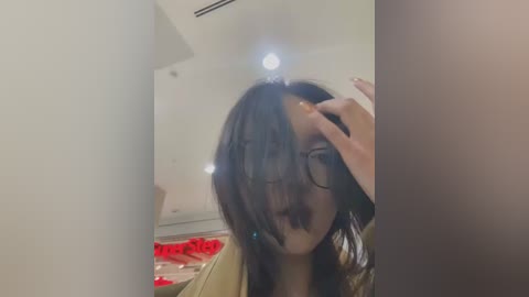 Media: Video of a young Asian woman with long dark hair, wearing glasses, adjusting her hair, in a modern, brightly-lit room with white walls and a red sign in the background.