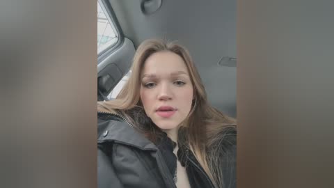 Media: Video of a young Caucasian woman with fair skin, light brown hair, and a neutral expression, wearing a black leather jacket and a white shirt. She is seated in the backseat of a car.